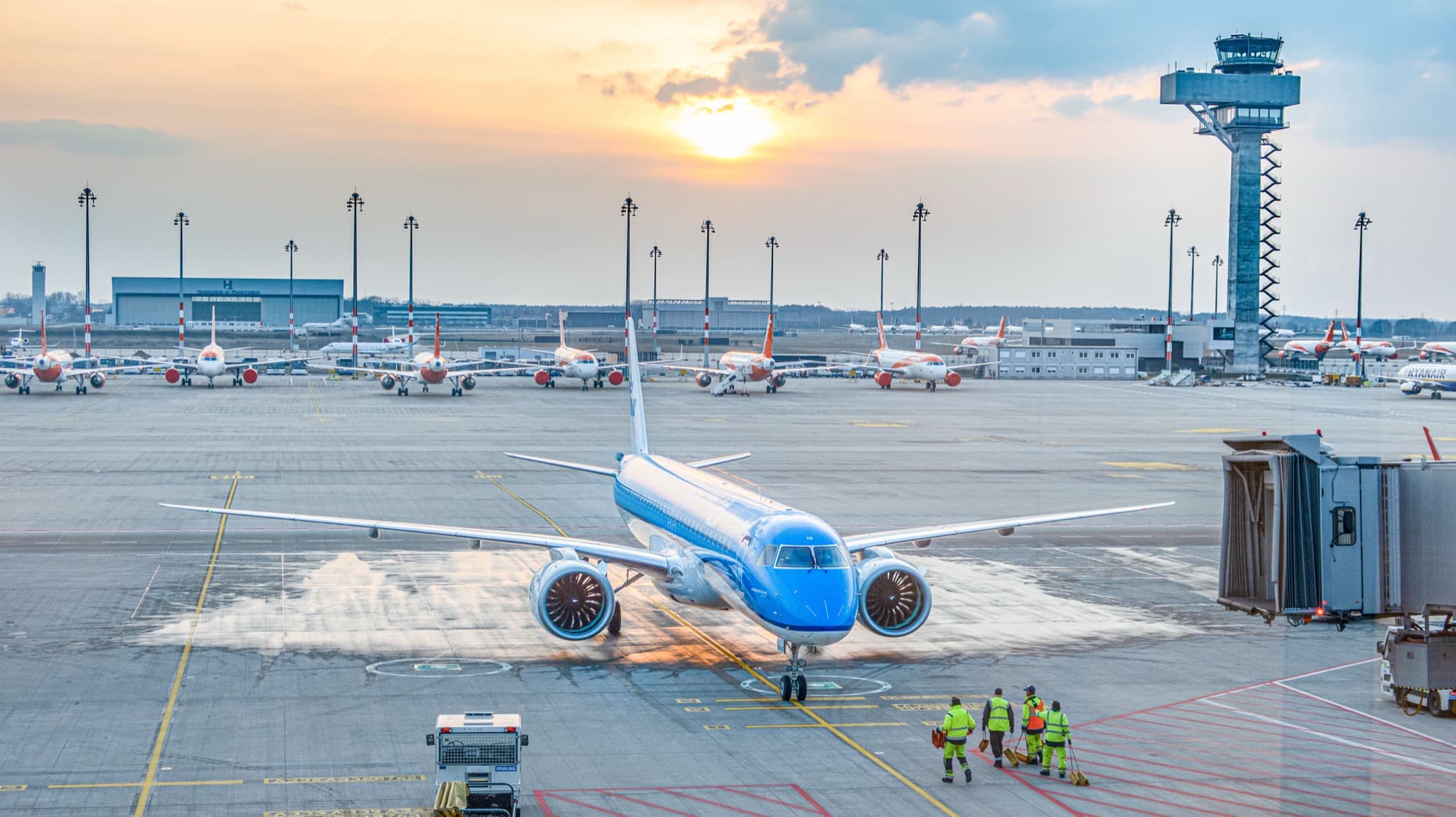 Avião Embraer E195-E2 taxia em pátio de aeroporto ao pôr do sol, cercado por outras aeronaves e equipe de solo (imagem de arquivo do modelo)
