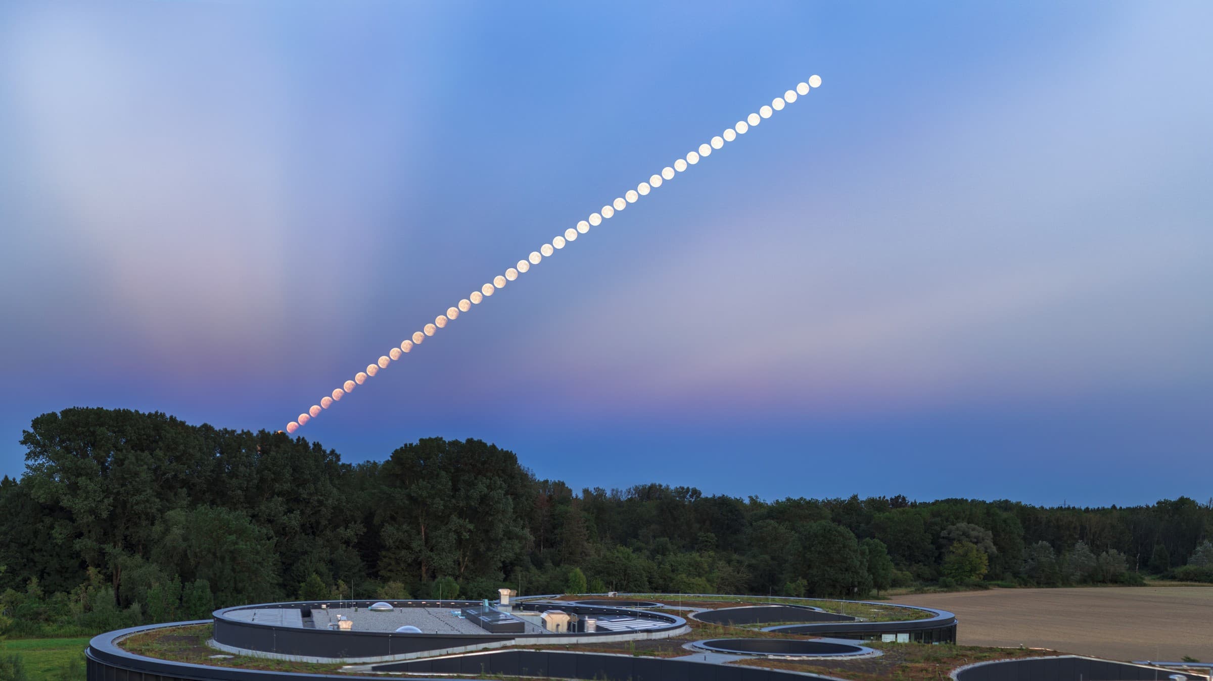 Sequência composta mostrando a Lua em eclipse subindo no céu ao anoitecer, sobre um prédio moderno