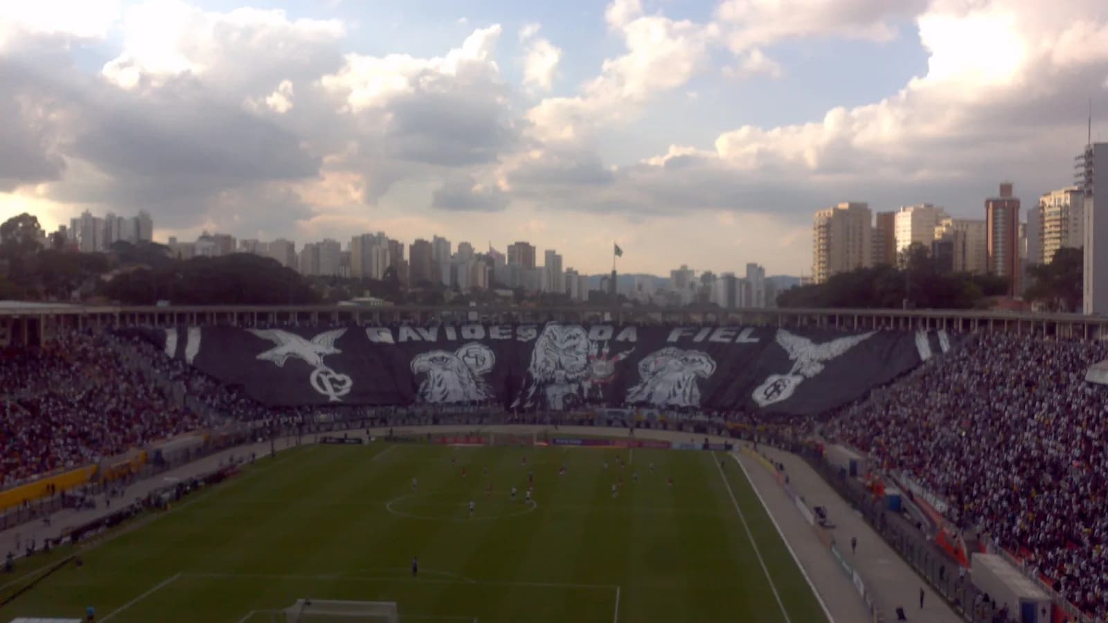 Torcida lotando arquibancada de estádio com mosaico em preto e branco durante jogo de futebol