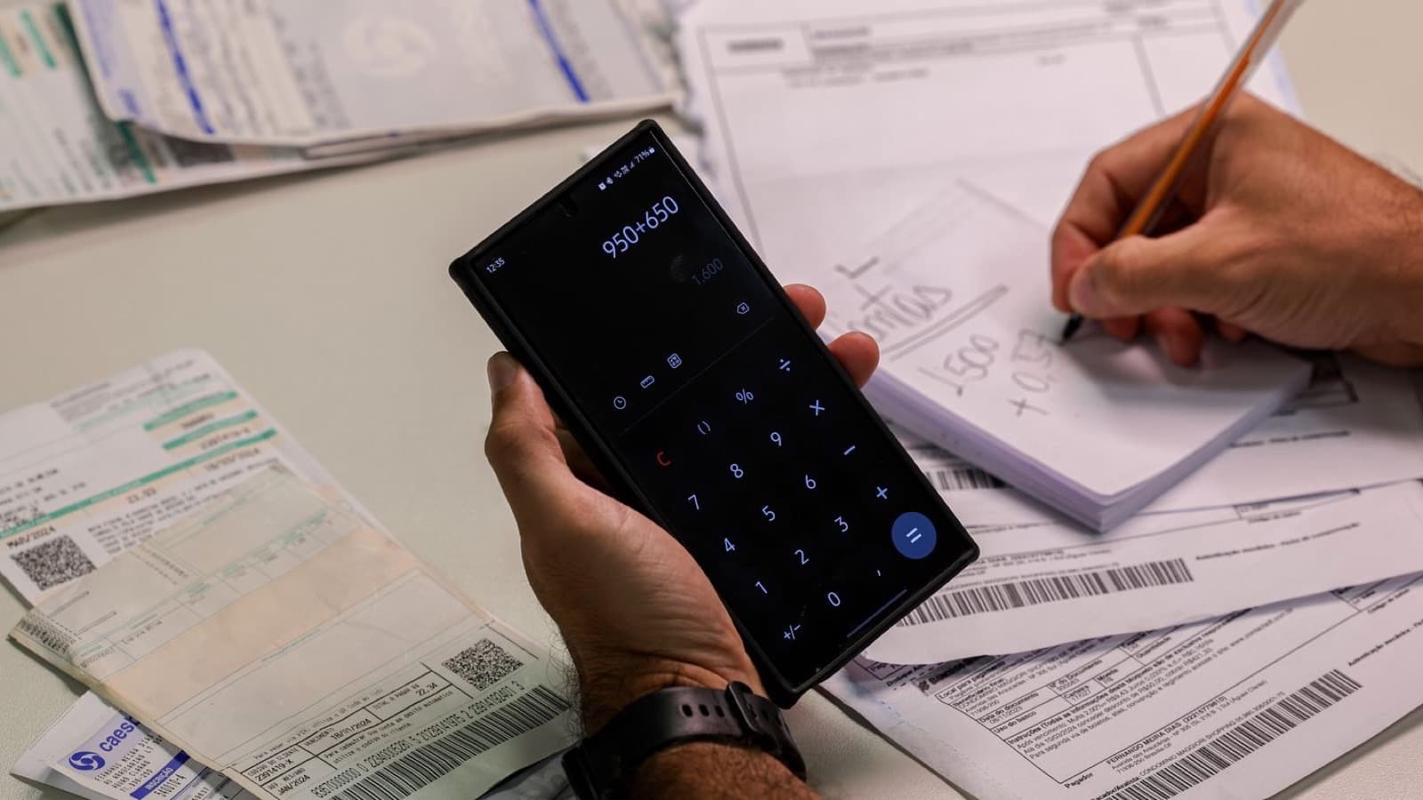 Mãos de uma pessoa segurando um celular com a calculadora aberta sobre uma mesa coberta de boletos e contas, enquanto outra mão anota somas em um bloco de papel, em imagem de arquivo