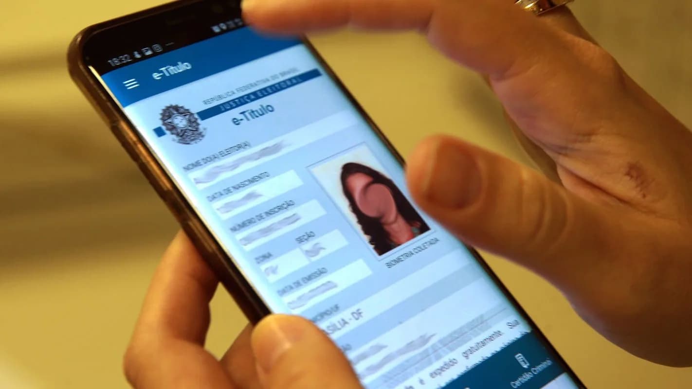 Mãos seguram um celular com o aplicativo e-Título aberto, mostrando os dados do título de eleitor