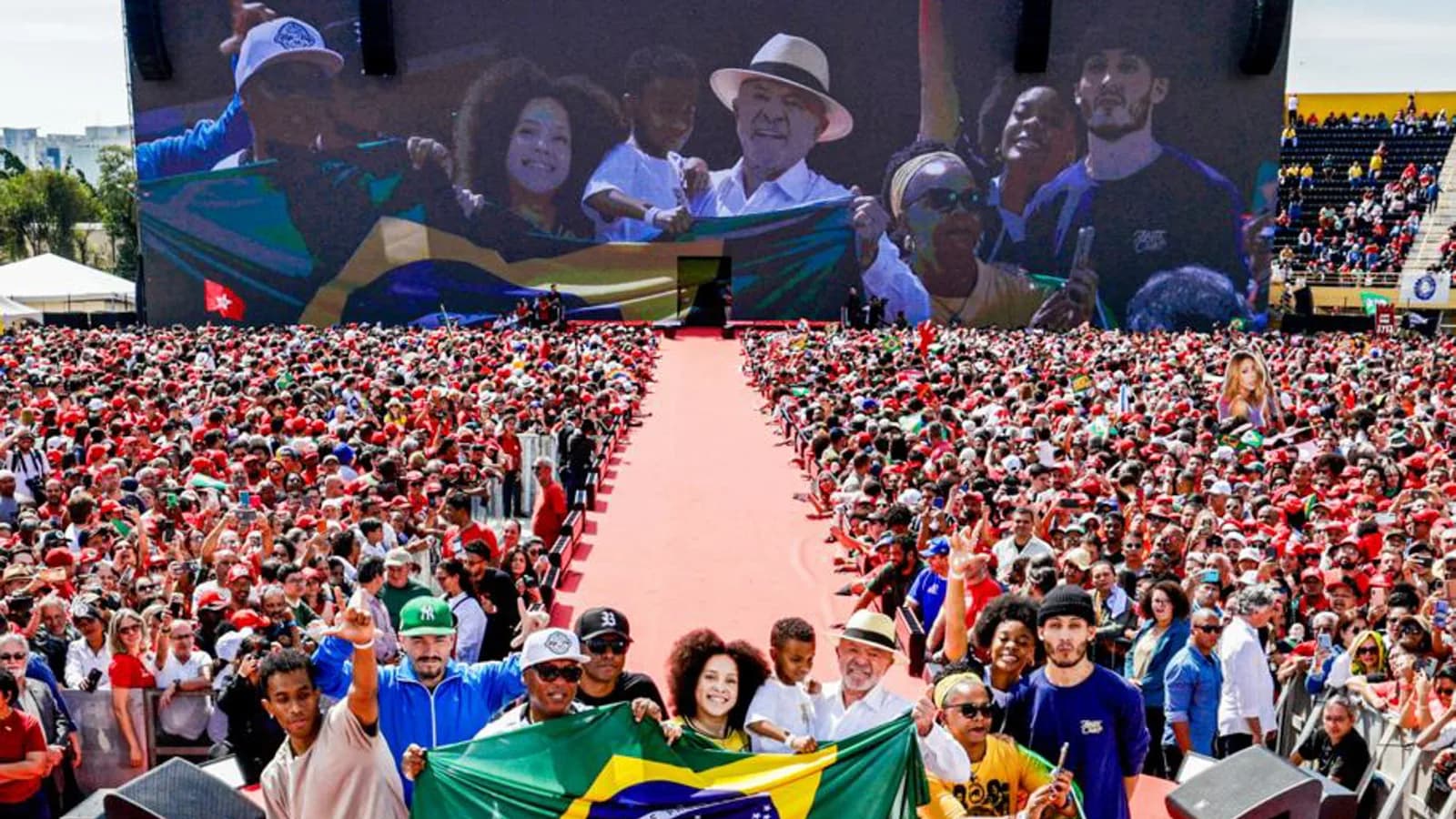 Apoiadores erguem bandeira do Brasil em frente a palanque com banner Presidente Lula Vice Alckmin no lançamento da campanha