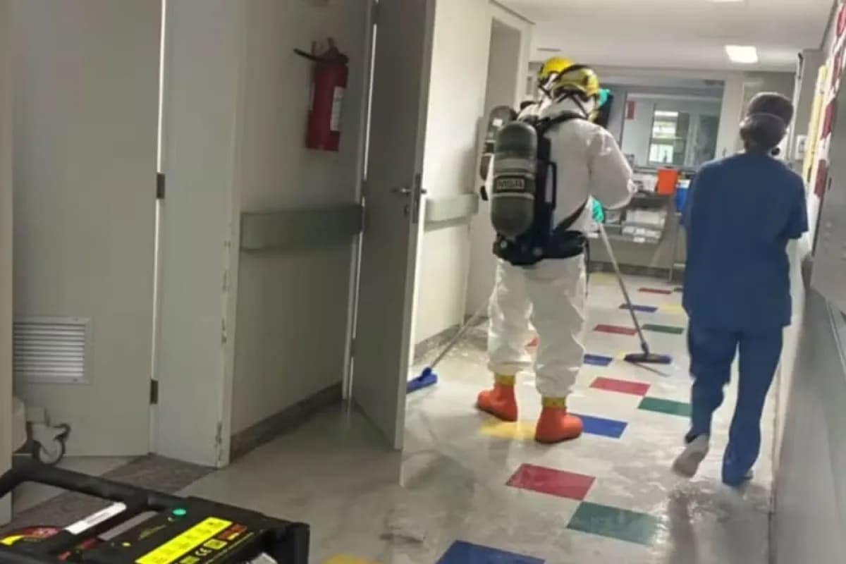 Bombeiro com traje de proteção e cilindro de oxigênio limpa corredor do CTI pediátrico da Santa Casa de BH após vazamento de ácido peracético, enquanto profissional de saúde se afasta ao fundo