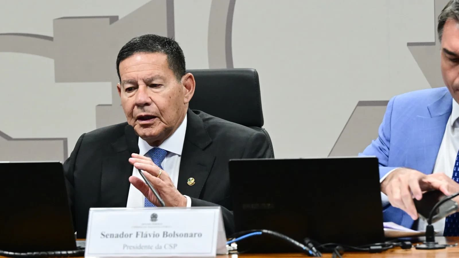 Senador Flávio Bolsonaro discursa sentado à mesa da Comissão de Segurança Pública do Senado, com placa de identificação como presidente da comissão