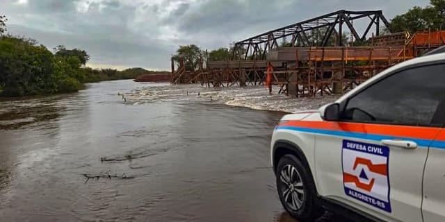 Viatura branca da Defesa Civil de Alegrete estacionada diante de estrada tomada pela água barrenta, ao lado de uma ponte metálica em obras sobre rio cheio, sob céu nublado