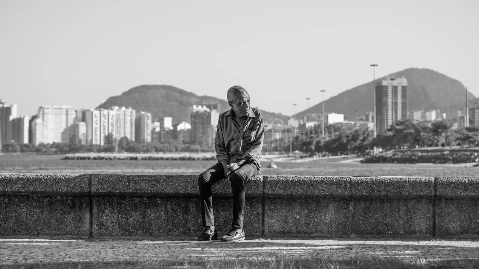 Homem idoso sentado sozinho em muro à beira-mar, com o Rio de Janeiro ao fundo
