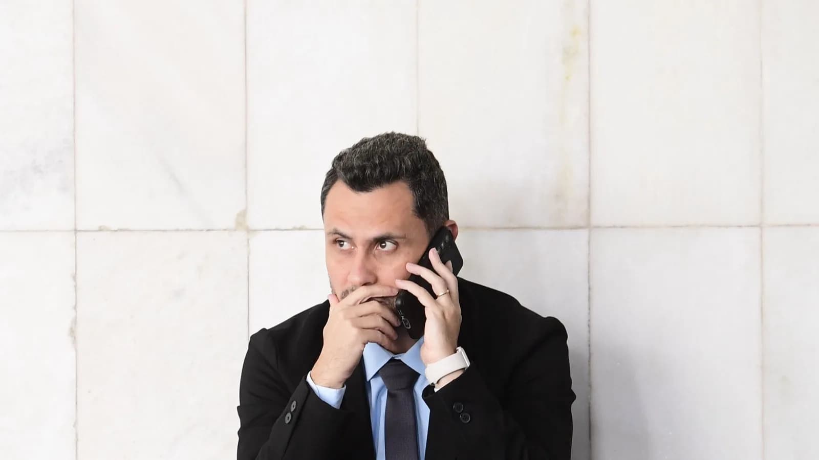 Senador Cleitinho, de terno preto, fala ao telefone celular agachado em corredor do Senado Federal