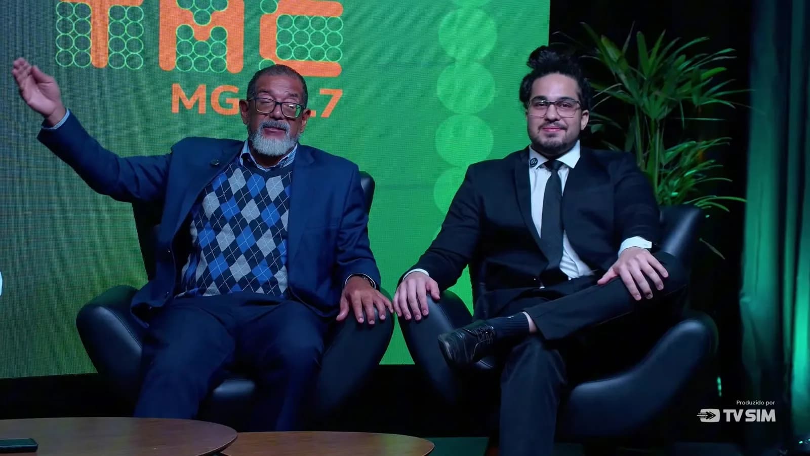 O superintendente regional do Trabalho em Minas Gerais, Carlos Calazans, sorri e gesticula com a mão erguida ao anunciar os acordos de escala 5x2 com Super Nosso e Vale, ao lado do advogado Igor Rodrigues, no programa A Cidade É Sua, da TMC