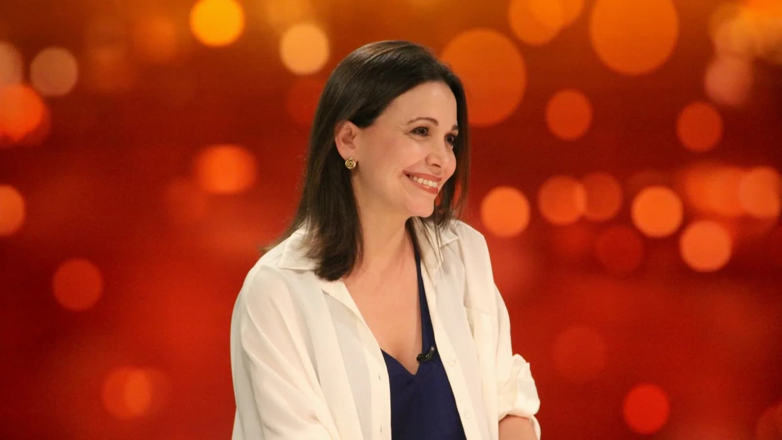 María Corina Machado sorri durante entrevista, com fundo desfocado em tons alaranjados