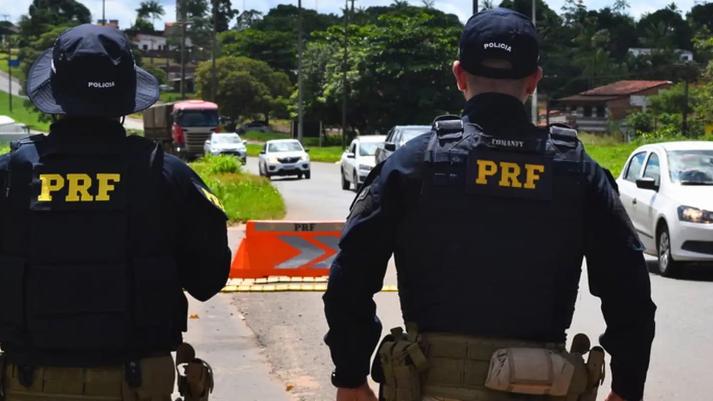 Dois policiais rodoviários federais de costas em fiscalização numa rodovia, com veículos ao fundo