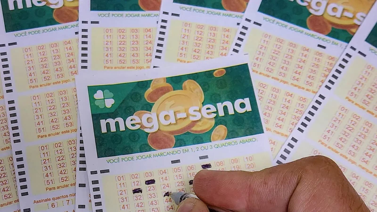 Volantes da Mega-Sena espalhados sobre uma mesa, com uma mão marcando dezenas com caneta; imagem de arquivo