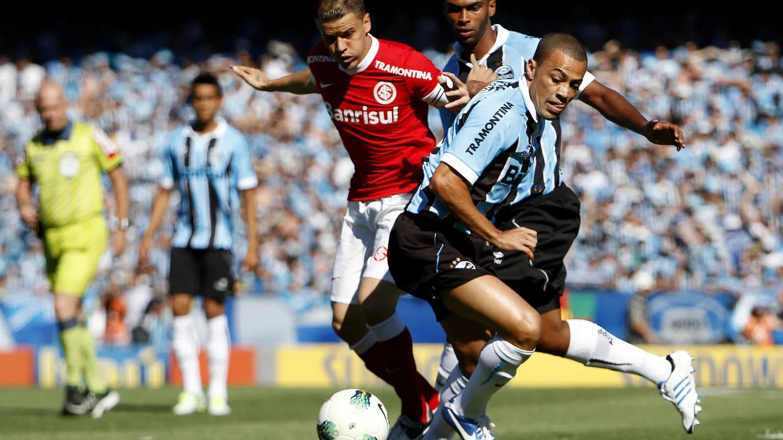 Jogador do Internacional disputa a bola com adversário do Grêmio em campo, com torcida ao fundo
