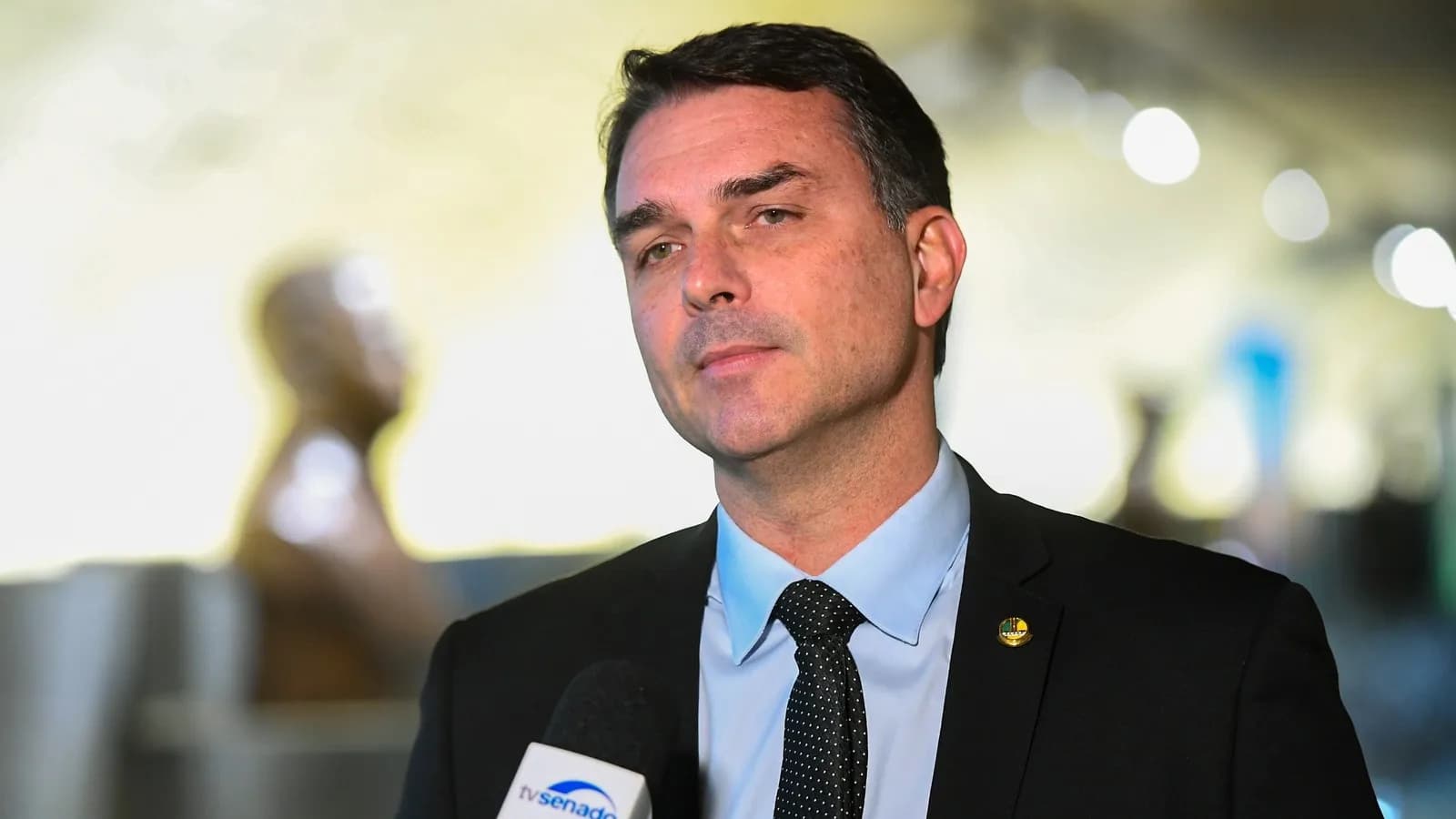 O senador Flávio Bolsonaro concede entrevista diante de um microfone da TV Senado, de terno escuro e camisa azul-clara