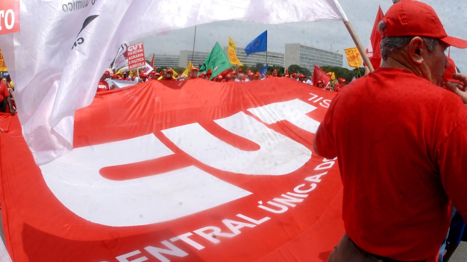 Sindicalistas seguram um bandeirão vermelho da CUT durante manifestação em Brasília; à direita, um homem de camisa e boné vermelhos aparece de costas, em imagem de arquivo