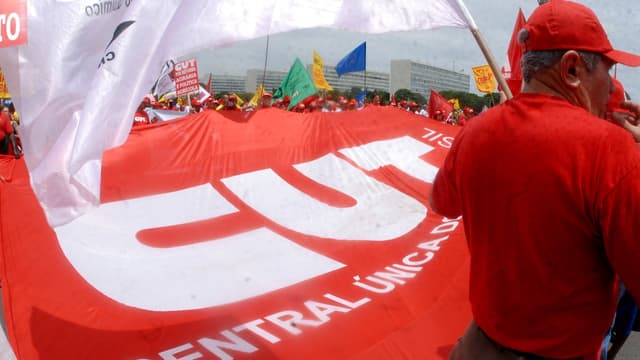 Sindicalistas seguram um bandeirão vermelho da CUT durante manifestação em Brasília; à direita, um homem de camisa e boné vermelhos aparece de costas, em imagem de arquivo