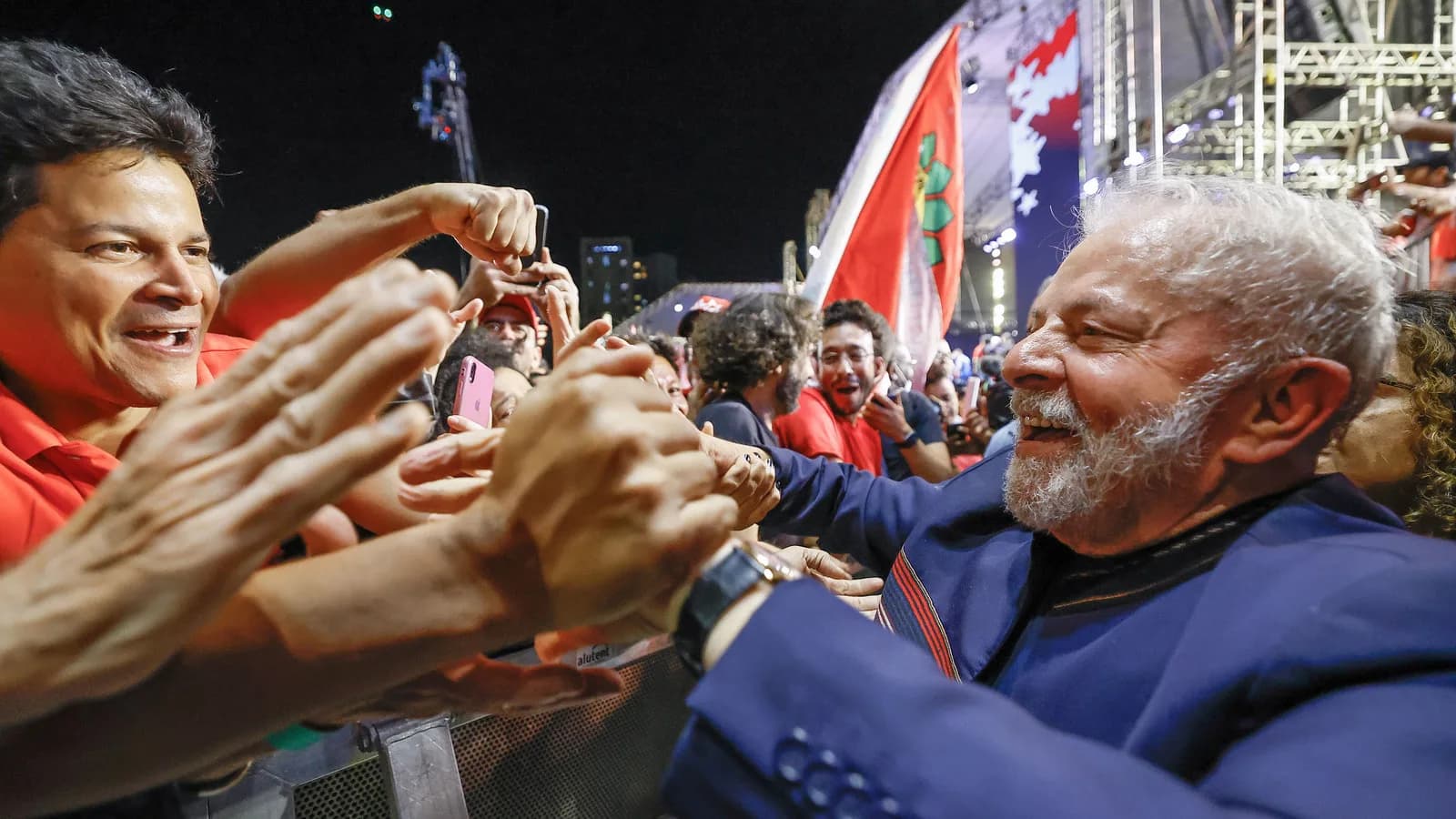 Lula sorrindo e cumprimentando apoiadores que estendem as mãos durante comício de campanha