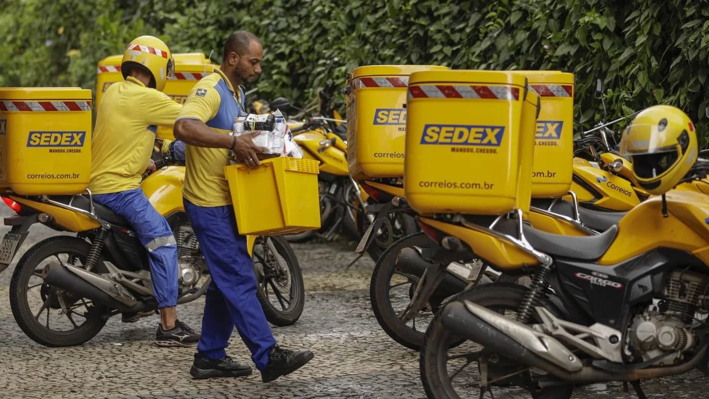 Carteiros dos Correios com uniforme amarelo carregam caixas ao lado de motos Sedex estacionadas
