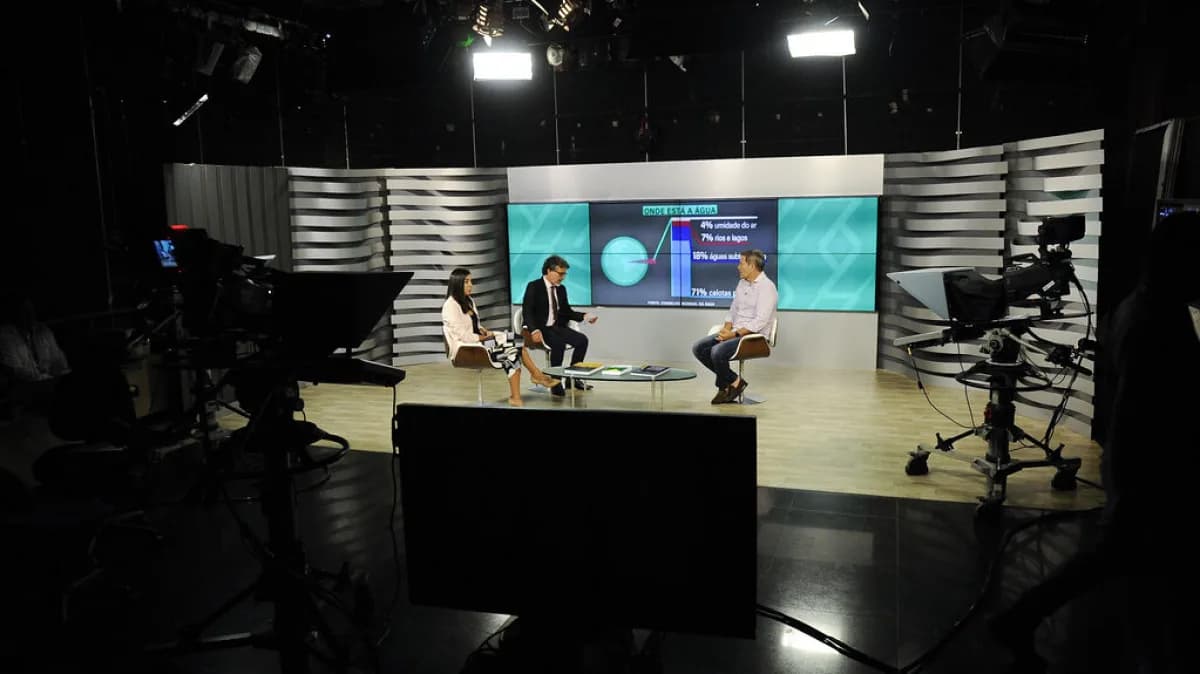 Estúdio de TV montado em formato de entrevista, com apresentador e dois entrevistados