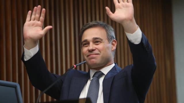 André Mendonça sorri com as mãos levantadas em evento oficial, de terno azul-marinho